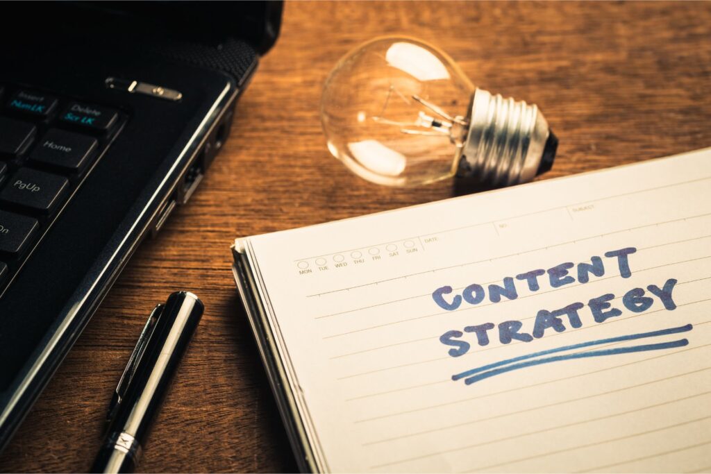 A notebook with the words "CONTENT STRATEGY" written on it, a pen, lightbulb, and laptop on a wooden surface symbolize content marketing