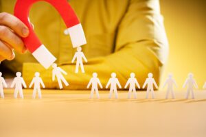 A hand holds a red and white magnet attracting paper cut-out people visualizing a strong lead magnet strategy.
