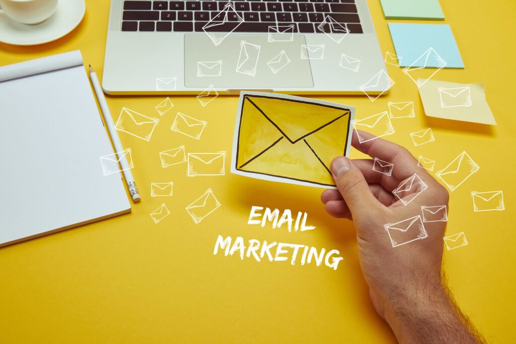 A hand holds a yellow envelope-shaped paper on a bright yellow background with a laptop, notepad, pencil, and scattered white envelope icons, illustrating email marketing trends.