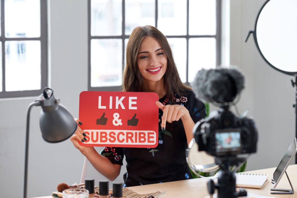 A young woman beauty influencer filming a polished video, holding a red "LIKE & SUBSCRIBE" sign, demonstrating YouTube marketing.