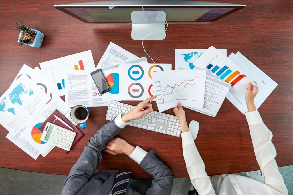 Two professionals at a cluttered desk analyze charts and data, illustrating efforts to track marketing campaign performance.