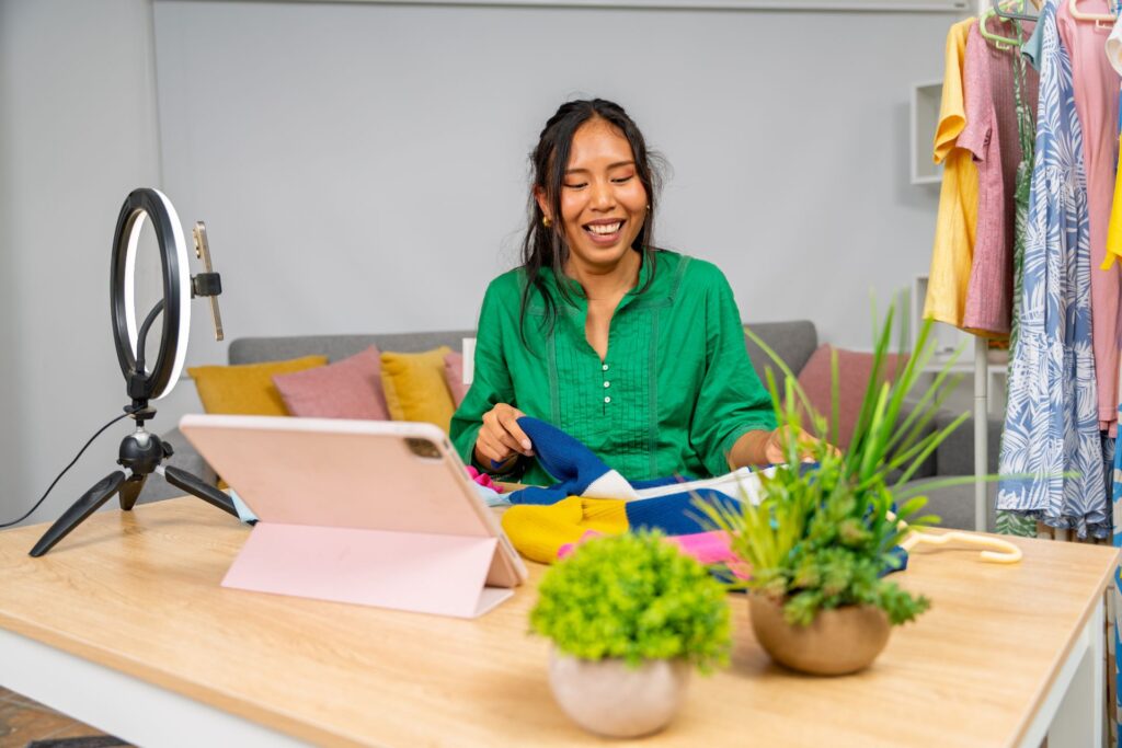 Smiling woman recording a tutorial with a tablet and ring light, using social media to amplify campaigns for her clothing business.