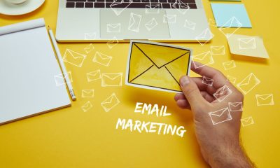 A hand holds a yellow envelope-shaped paper on a bright yellow background with a laptop, notepad, pencil, and scattered white envelope icons, illustrating email marketing trends.