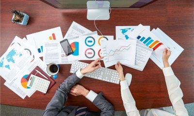 Two professionals at a cluttered desk analyze charts and data, illustrating efforts to track marketing campaign performance.