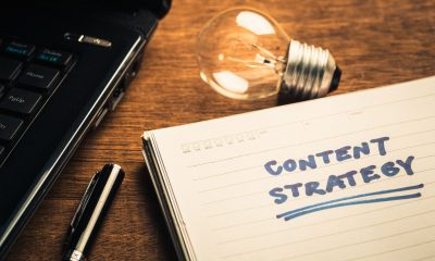 A notebook with the words "CONTENT STRATEGY" written on it, a pen, lightbulb, and laptop on a wooden surface symbolize content marketing