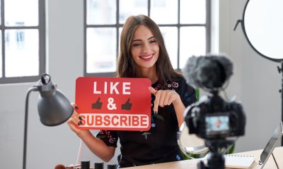 A young woman beauty influencer filming a polished video, holding a red "LIKE & SUBSCRIBE" sign, demonstrating YouTube marketing.